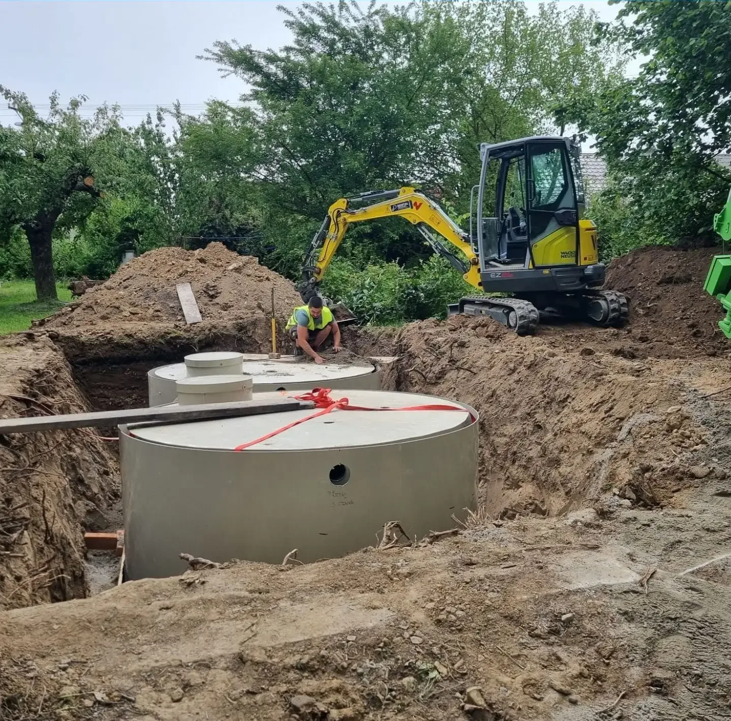 Minibagr osazuje velké betonové dešťové nádrže do výkopu na zahradě