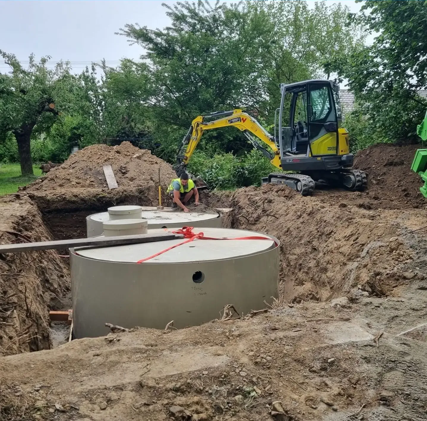 Minibagr osazuje velké betonové dešťové nádrže do výkopu na zahradě