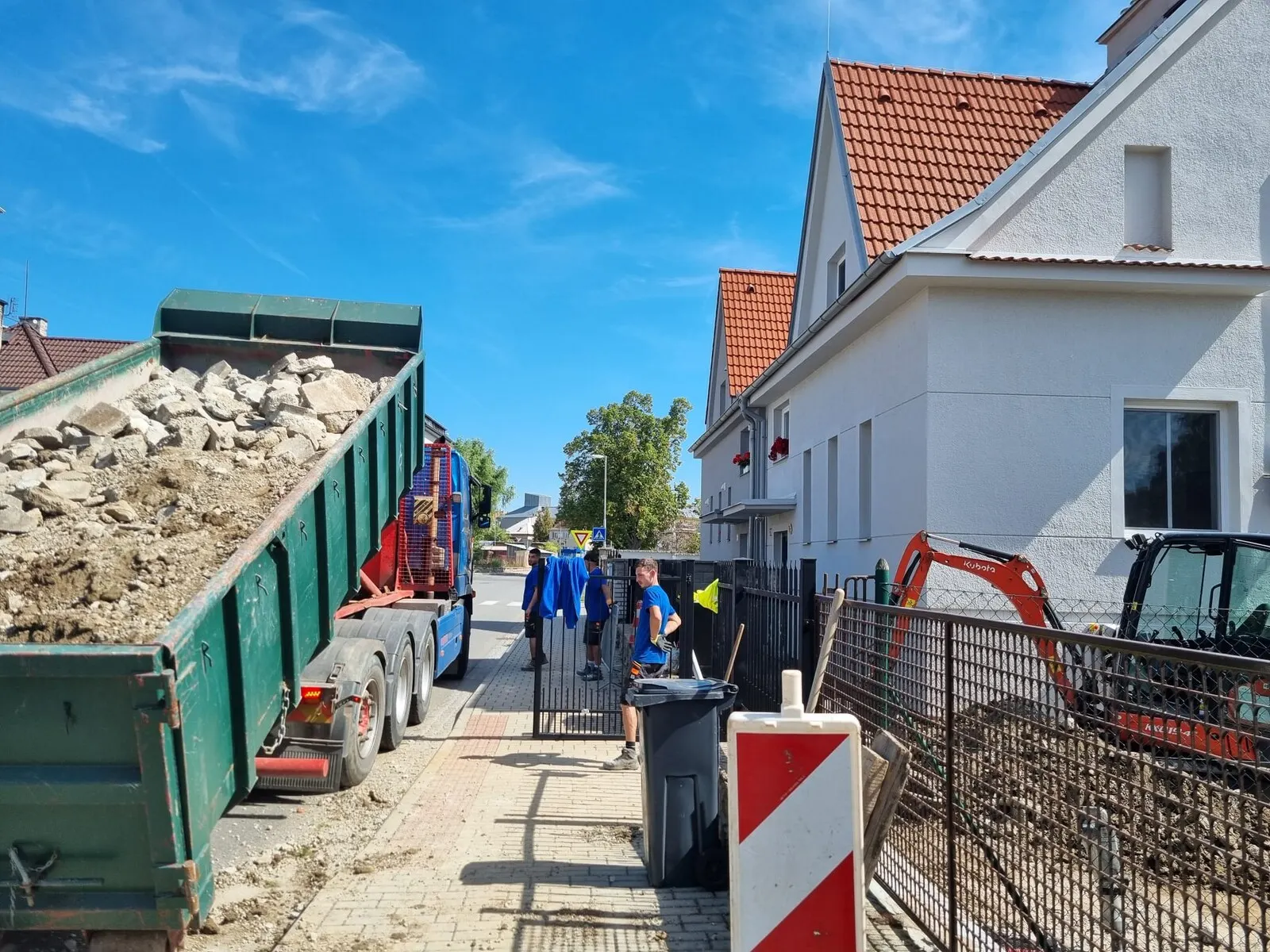 Nákladní auto s kontejnerem na suť a minibagr v ulici u rekonstruovaného domu